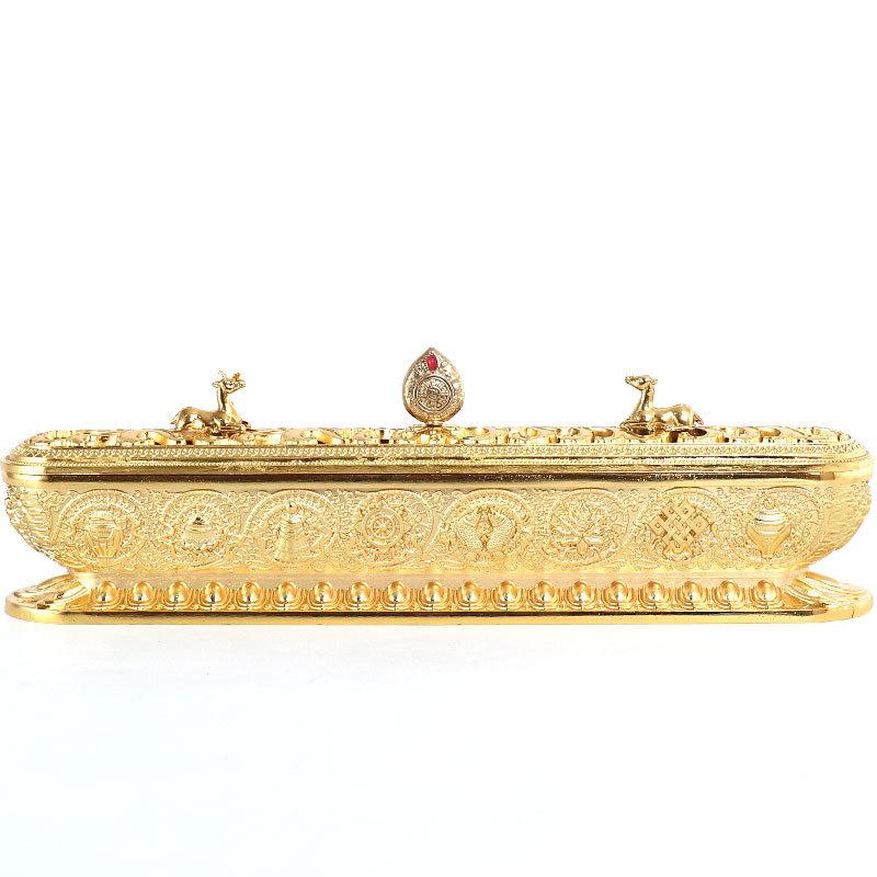 Gold-tone Tibetan incense burner with elongated rectangular body, decorative lid, and Buddhist motifs on white
